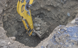 A yellow excavator is shown digging a deep hole preparation for underground pipe rehabilitation