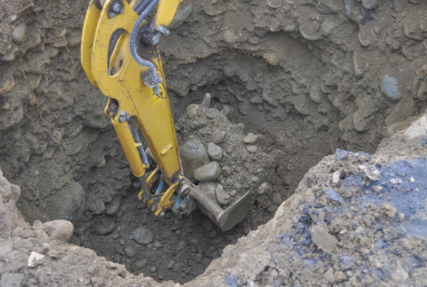 A yellow excavator is shown digging a deep hole preparation for underground pipe rehabilitation