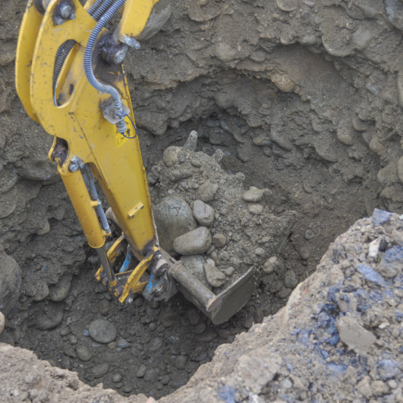 A yellow excavator is shown digging a deep hole preparation for underground pipe rehabilitation