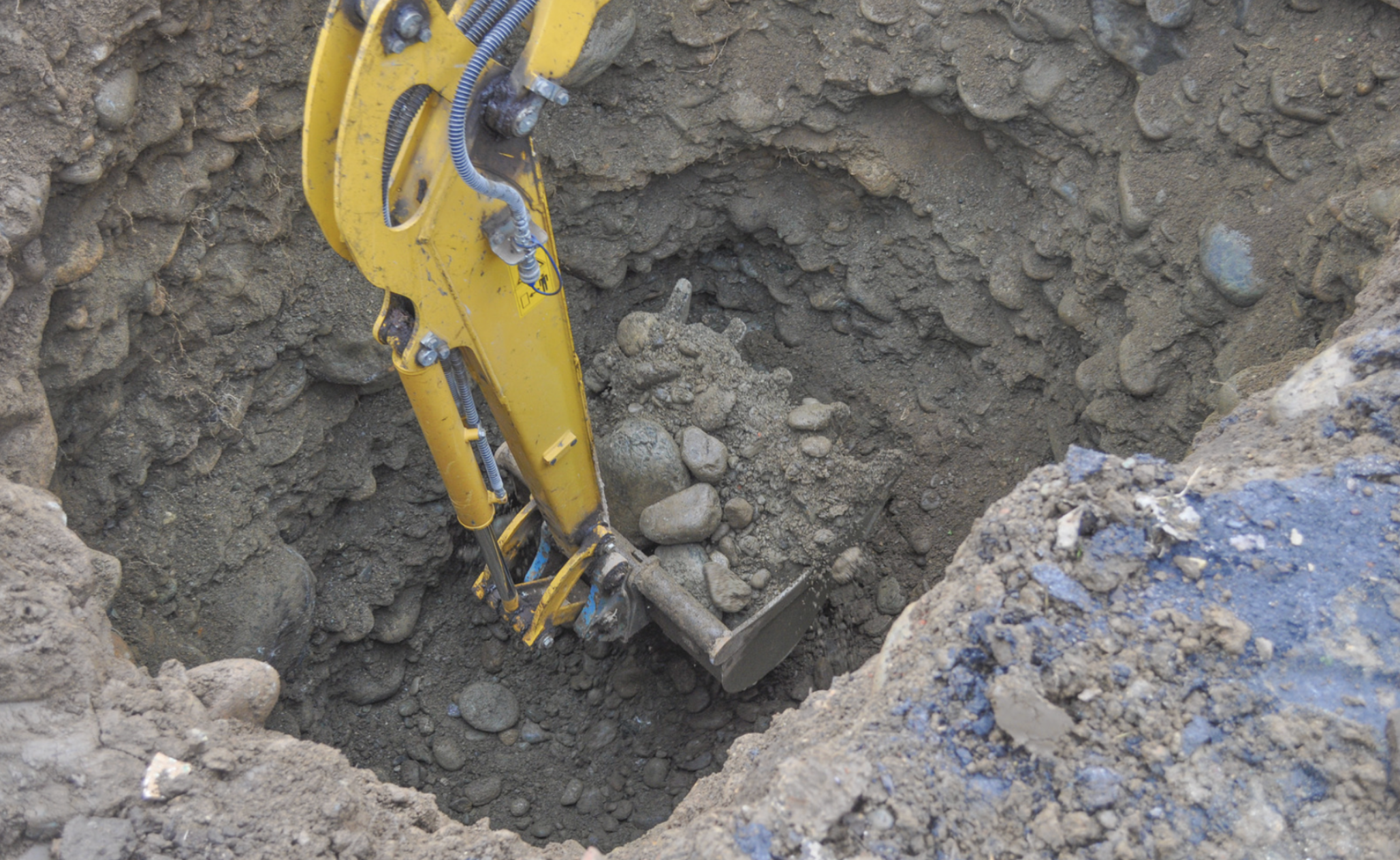 A yellow excavator is shown digging a deep hole preparation for underground pipe rehabilitation